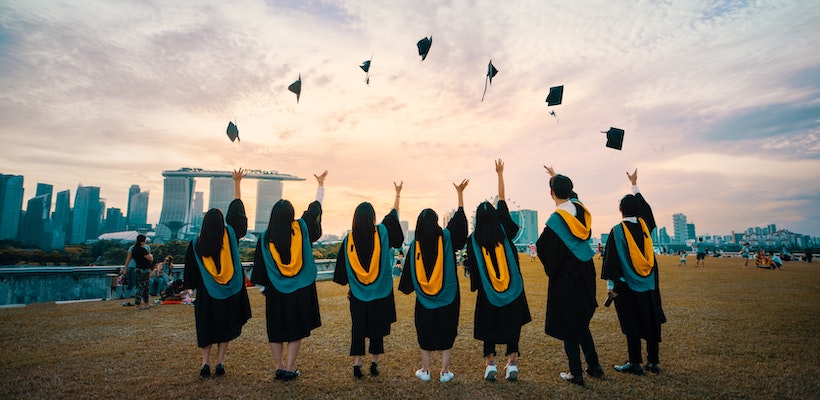 A group of 7 college graduates face away from the camera, stand in a line and toss their caps up forming an arch above their heads.