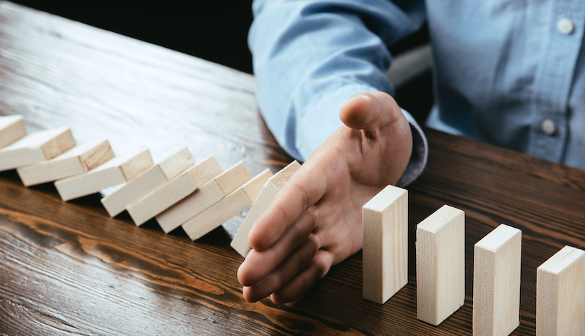 A man sits at a table with blocks in a line. His hand intersects a line of block stopping the flow of blocks cascading, implying sequence risk protection.